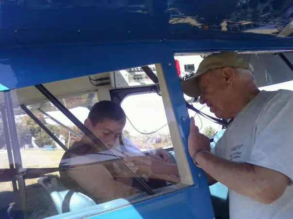 Bobby in the cockpit of a blue Citabria while Billy leans in to fasten his harness