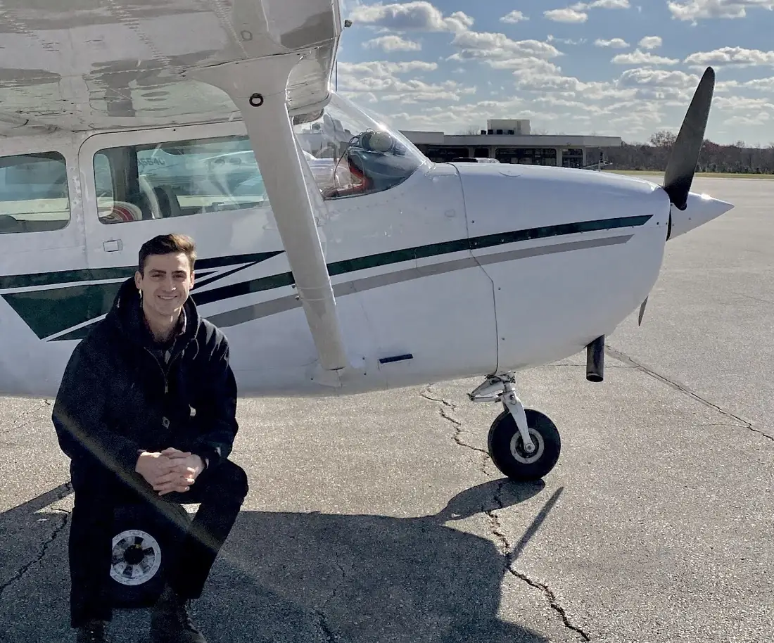 A man kneeling beside a white Cessna on an airport ramp under a blue sky