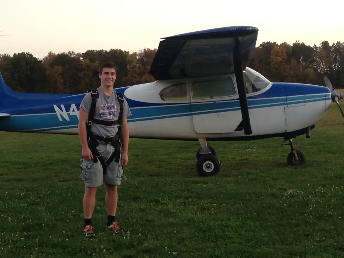 A young man in a parachute harness standing in front of a blue and white airplane on grass